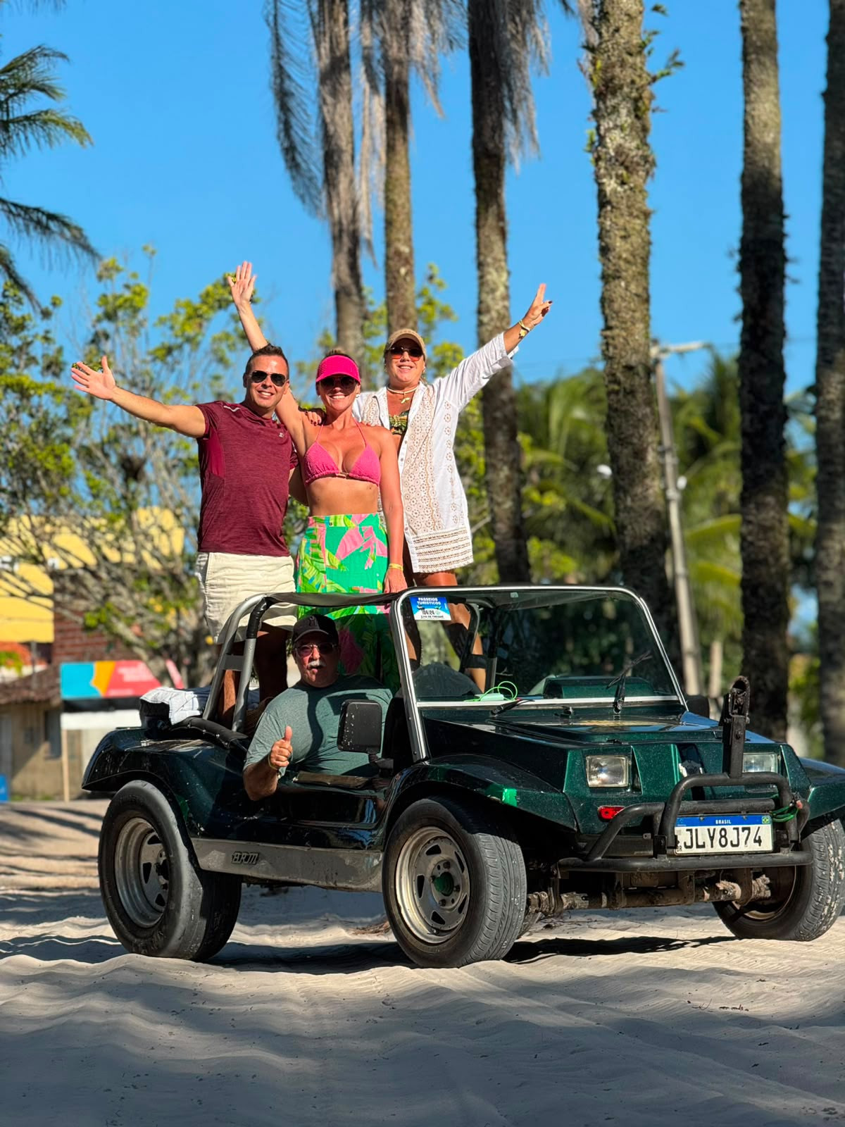 Grupo comemorando em pé em um buggy verde numa rua de areia entre coqueiros